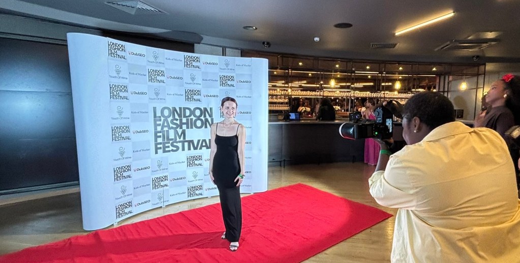 A woman posing on a red carpet at the London Fashion Film Festival, with a backdrop featuring the festival's logo. A photographer is capturing the moment.